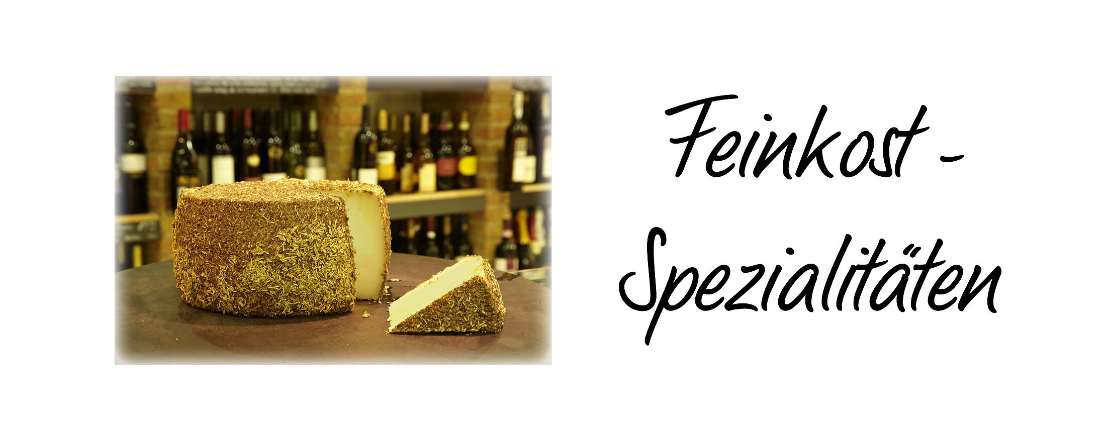 Ein großer Laib Rosmarinmanchego, angeschnitten auf einem Tisch, im Hintergrund ein Weinregal & die Aufschrift "Feinkost"