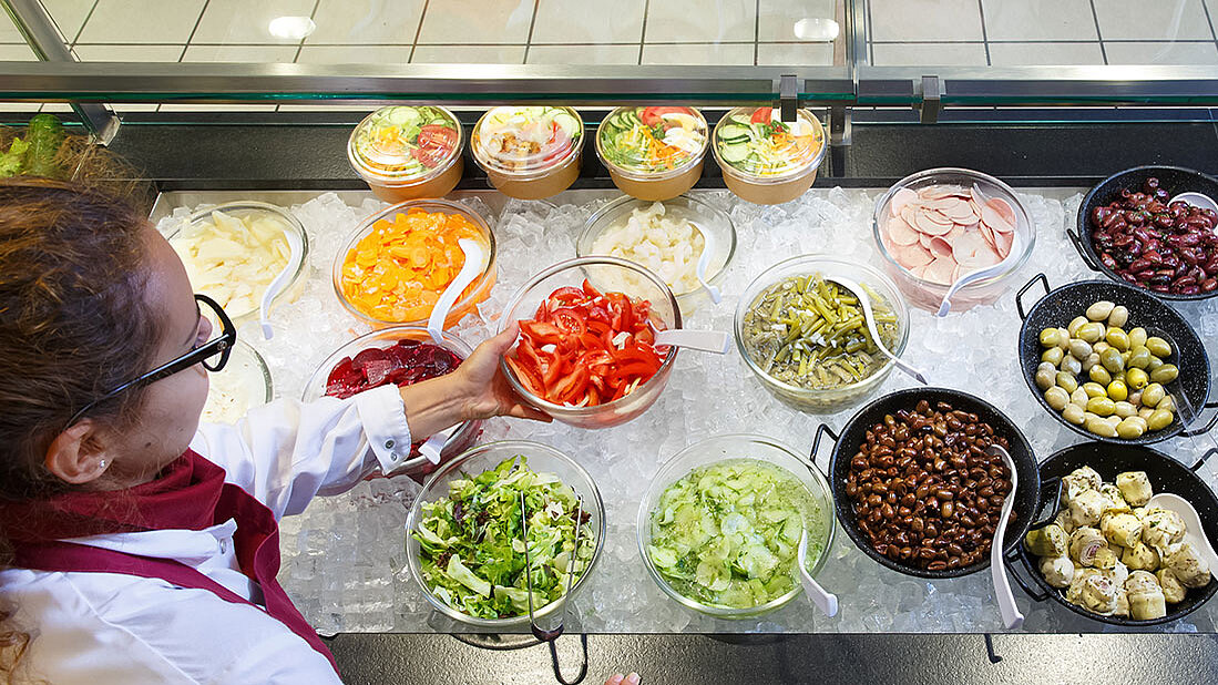 Verkäuferin greift in eine Salattheke mit vielen frischen Feinkostsalaten, Antipasti, Oliven & eingelegtem Gemüse auf Eis