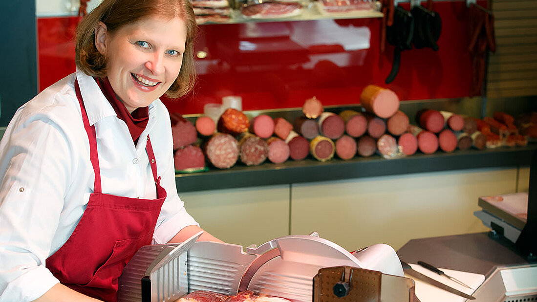 Alexandra Schmit in roter Schürze bedient eine Aufschnittmaschine vor einer Theke mit vielfältiger Wurst- & Fleischware