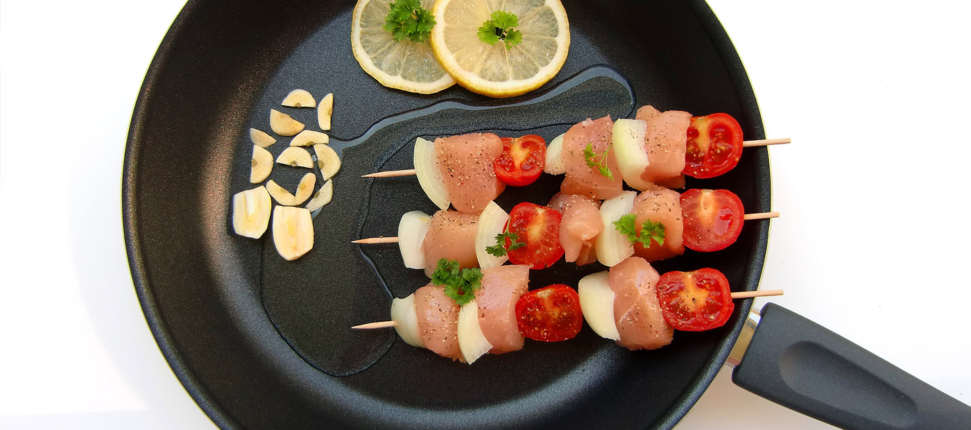 Zwei Fleischspieße mit Zwiebeln, Tomaten & Petersilie in einer Pfanne, daneben Knoblauchstücke & Zitronenscheiben